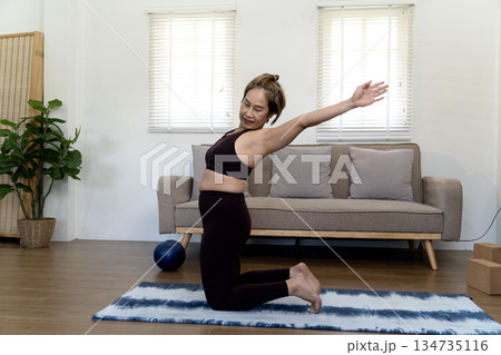 Adult woman 50+ in workout clothes kneeling on a mat and stretching Adult woman 50+ in workout clothes kneeling on a mat and stretching 134735116
