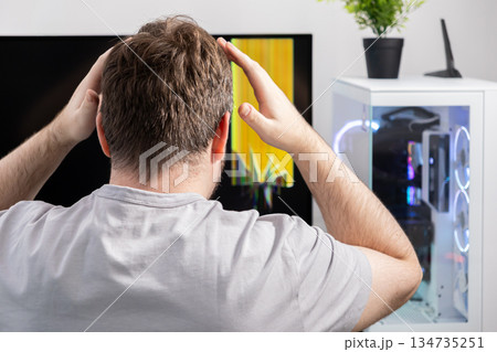 Frustrated man clutching head, gazing at a desktop computer monitor showing severe glitch errors, symbolizing technical malfunction and urgent repair need 134735251