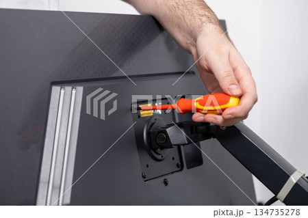 Person's hand uses a precision screwdriver to securely fasten a monitor arm onto a computer display, illustrating practical home office equipment assembly and installation 134735278