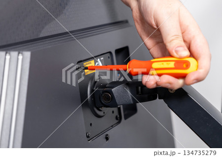 Persons hand using a bright orange screwdriver to secure a computer monitor mount screw, illustrating careful technical assembly and ergonomic workspace setup Persons hand using a bright orange screwdriver to secure a computer monitor mount screw, illustrating careful technical assembly and ergonomic workspace setup 134735279