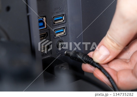 User's hand plugging a black DC-IN power cable into the back panel of a modern electronic device, illustrating essential connectivity and setup 134735282