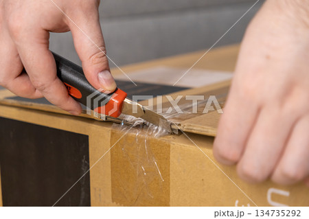 Hand uses utility knife to cut clear tape on brown cardboard delivery box, symbolizing modern e-commerce and new product unpacking Hand uses utility knife to cut clear tape on brown cardboard delivery box, symbolizing modern e-commerce and new product unpacking 134735292