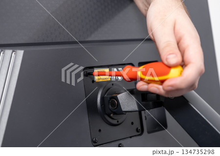 Hand holding a colorful screwdriver to secure a screw on a black monitor mount with a visible WARNING label, representing assembly and technical maintenance Hand holding a colorful screwdriver to secure a screw on a black monitor mount with a visible WARNING label, representing assembly and technical maintenance 134735298