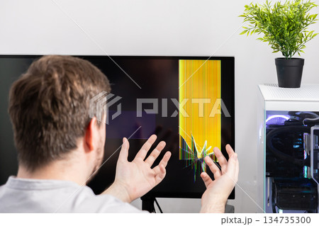 Frustrated person with hands reacting to a severely broken computer monitor, displaying colorful lines, signifying technical damage, repair, and unexpected expense Frustrated person with hands reacting to a severely broken computer monitor, displaying colorful lines, signifying technical damage, repair, and unexpected expense 134735300