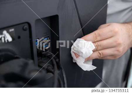 Persons hand meticulously wiping dust from the rear panel of a black computer monitor using a disposable wipe, ensuring electronics cleanliness and workspace hygiene 134735311