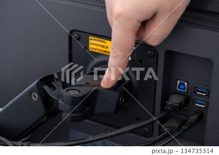 Hand adjusting a VESA monitor arm mount on a computer screen, visible warning label and cables, demonstrating ergonomic desk setup Hand adjusting a VESA monitor arm mount on a computer screen, visible warning label and cables, demonstrating ergonomic desk setup 134735314