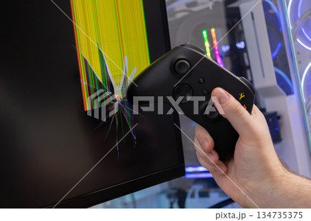 Player's hand grasps a black video game controller, hitting a broken computer monitor with colorful distorted pixels, signifying intense gaming rage and equipment failure Player's hand grasps a black video game controller, hitting a broken computer monitor with colorful distorted pixels, signifying intense gaming rage and equipment failure 134735375