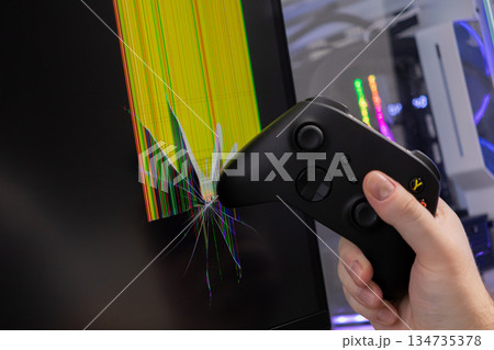 Hand holding a black game controller smashing into a computer monitor, causing widespread screen cracks and illustrating intense gaming frustration Hand holding a black game controller smashing into a computer monitor, causing widespread screen cracks and illustrating intense gaming frustration 134735378