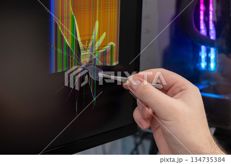 A person's hand uses tweezers to meticulously remove a tiny glass shard from a severely cracked computer monitor, demonstrating electronic device screen repair 134735384