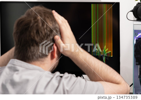 Man reacting with frustration to a severely broken computer monitor displaying vibrant vertical lines and a visible crack, symbolizing sudden technological failure 134735389