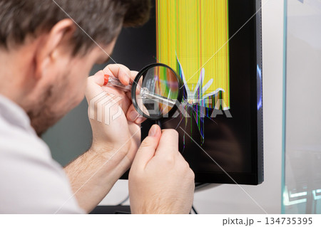 Technician examines cracked computer monitor with magnifying glass and screwdriver, symbolizing electronics repair and technical diagnostics 134735395