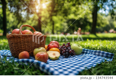 Wicker basket filled with fresh apples, grapes, and peaches placed on checkered tablecloth in sunlit autumn forest, symbolizing seasonal fruit harvest. 134743544