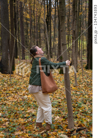 Woman in a green jacket shakes a tree in an autumn forest, causing yellow leaves to fall, playful and joyful seasonal moment Woman in a green jacket shakes a tree in an autumn forest, causing yellow leaves to fall, playful and joyful seasonal moment 134743893