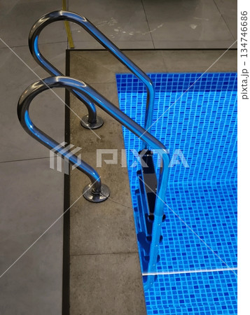 Stairs and handrails in a modern swimming pool with bright blue mosaic tiles and lighting Stairs and handrails in a modern swimming pool with bright blue mosaic tiles and lighting 134746686