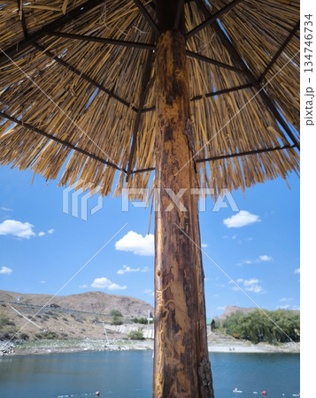 A view from under a thatched beach umbrella of a lake and hills on a sunny day 134746734
