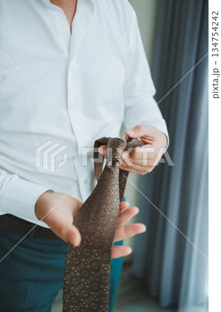 A Man Thoughtfully Preparing to Tie a Brown Designer Tie in a Stylish and Elegant Setting A Man Thoughtfully Preparing to Tie a Brown Designer Tie in a Stylish and Elegant Setting 134754242