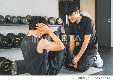 スポーツジムで腹筋・腹斜筋を鍛える・筋トレをする男性と指導するパーソナルトレーナー 134754548