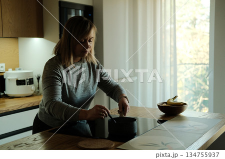 Woman stands in modern kitchen holding spoon while looking down at small pot on induction stove. Solo dining trends, independent living, cooking for one. Woman stands in modern kitchen holding spoon while looking down at small pot on induction stove. Solo dining trends, independent living, cooking for one. 134755937