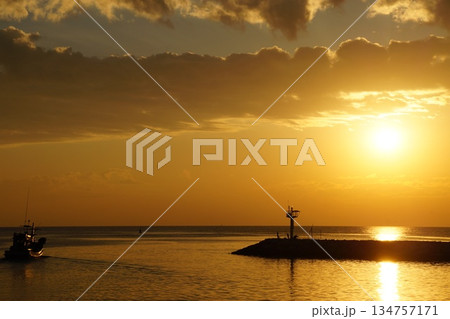 A large, bright, golden sun rises from the horizon against a backdrop of a stone pier, large rocks, and stalks of thatched grass. A golden path from the rising sun lays across the surface of the gentl A large, bright, golden sun rises from the horizon against a backdrop of a stone pier, large rocks, and stalks of thatched grass. A golden path from the rising sun lays across the surface of the gentl 134757171