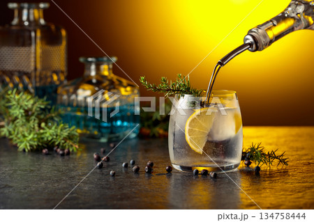 Gin is poured from a bottle into a glass with ice and lemon slice. Gin is poured from a bottle into a glass with ice and lemon slice. 134758444