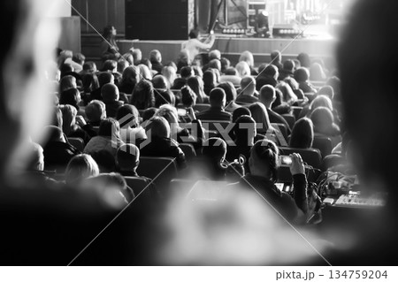Black and white shot of an audience sitting in a theater or concert hall, looking at a brightly lit stage. A person in the foreground is filming the performance on a smartphone Black and white shot of an audience sitting in a theater or concert hall, looking at a brightly lit stage. A person in the foreground is filming the performance on a smartphone 134759204