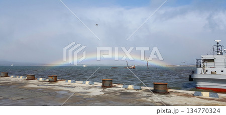 Post-storm scene in a Kuril Islands bay. A vibrant rainbow arcs over a pier, mooring, a docked ship, and a distant shipwreck. 134770324
