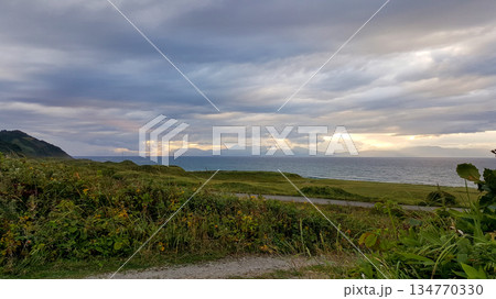 Evening coastal view on the Kuril Islands. Rolling green hills meet the sea under a dramatic sky. Sunlight streams through the clouds 134770330