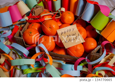 Box with fresh tangerines, festive colorful garlands and sticky note with handwritten phrase Happy new 2026. Message with congratulations and celebration mood of winter holidays. 134770665