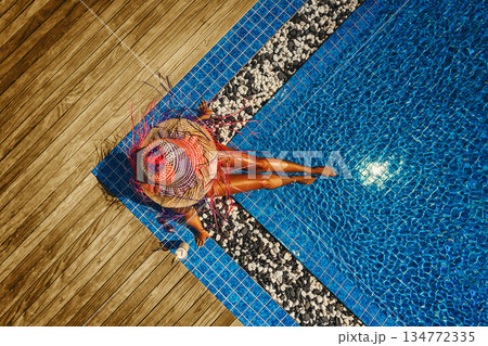 Drone view of a woman in a colorful straw hat relaxing by an infinity pool on a wooden deck witj coctail 134772335