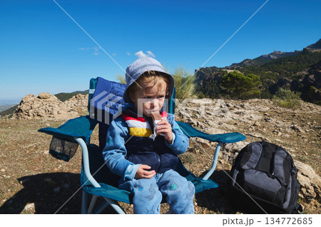 Young Child Eating Snack In Blue Camping Chair On Mountain Trail With Backpack Nearby Young Child Eating Snack In Blue Camping Chair On Mountain Trail With Backpack Nearby 134772685