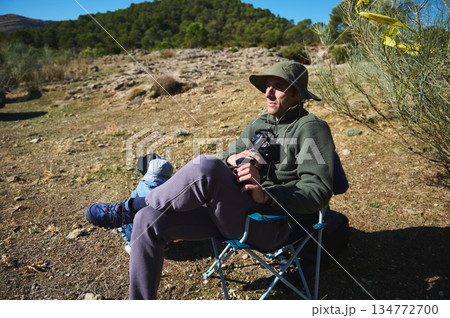 Relaxed Photographer Sitting in Camping Chair Outdoors With Camera in Sunny Natural Setting Relaxed Photographer Sitting in Camping Chair Outdoors With Camera in Sunny Natural Setting 134772700