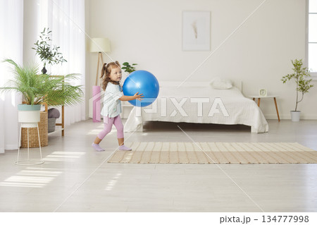 Little cute child girl holding fit ball in hands standing in living room at home Little cute child girl holding fit ball in hands standing in living room at home 134777998