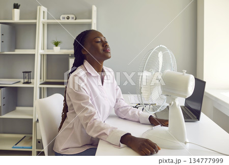 Young african american woman working at office using electric fan during heatwave in office. 134777999
