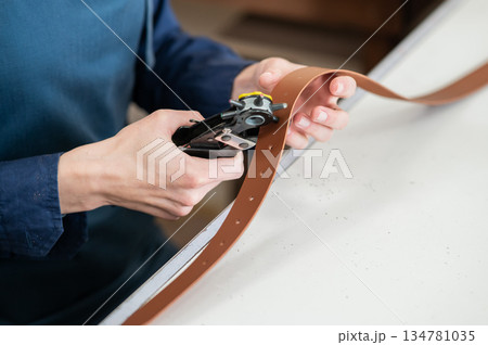 A woman makes holes in a leather belt. Leatherworker's workshop.  134781035