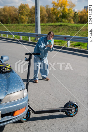 Woman examines and photographs the damage caused by a collision between an electric scooter and a car. 134781071