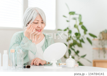 家で鏡を見ながらメイク,アイメイクをするグレーヘアーのシニア女性 134784141