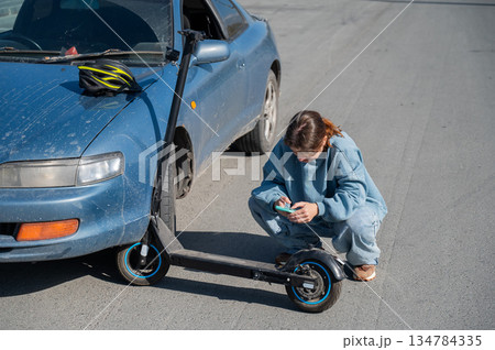 Woman examines and photographs the damage caused by a collision between an electric scooter and a car. 134784335