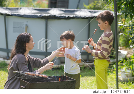 Caucasian woman grilling sausages on a barbecue with her two sons. Caucasian woman grilling sausages on a barbecue with her two sons. 134784634