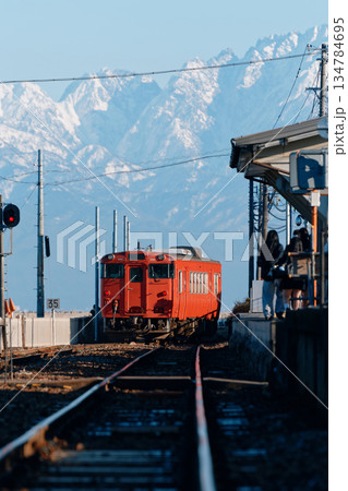 雪山に向かって走る橙色のローカル線とそれを見送る人々 134784695