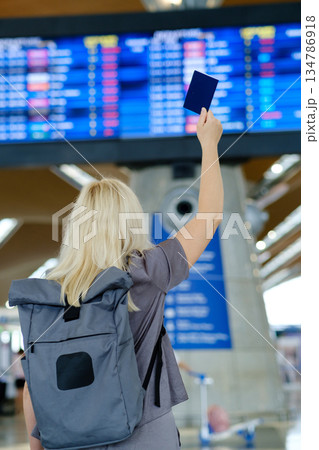 Traveler with a backpack raises a passport at the airport while checking flight information on a digital display board 134786918