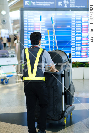 Airport staff member performs cleaning duties with a cart in busy terminal during afternoon hours as travelers move about 134786921