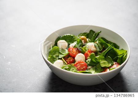 Salad with cherry tomatoes, spinach leaves and mozzarella balls in white bowl on terrazzo surface with copy space 134786965