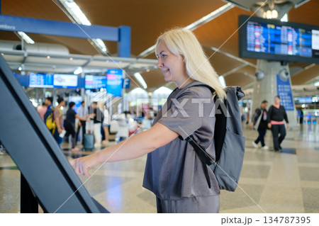 Woman using a touchscreen information kiosk at an airport terminal while travelers wait in the busy background during the daytime Woman using a touchscreen information kiosk at an airport terminal while travelers wait in the busy background during the daytime 134787395