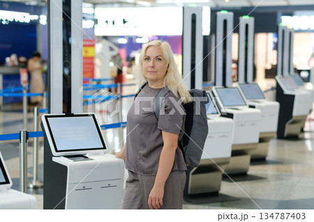 Woman with backpack interacting with check-in kiosk in modern airport terminal during busy travel season Woman with backpack interacting with check-in kiosk in modern airport terminal during busy travel season 134787403