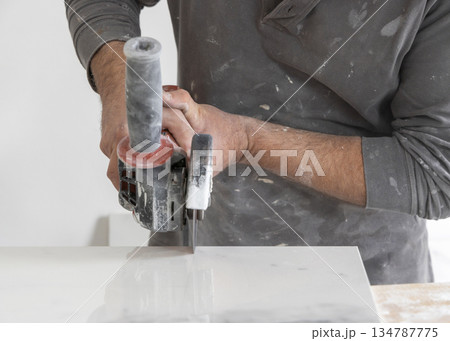 Craftman in grey shirt cuts a gray marble tile using an angle grinder. Handy men at work, hands closeup Craftman in grey shirt cuts a gray marble tile using an angle grinder. Handy men at work, hands closeup 134787775