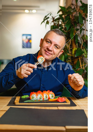 Cheerful man poses with a sushi roll over a neatly arranged sushi platter. Cheerful man poses with a sushi roll over a neatly arranged sushi platter. 134789078