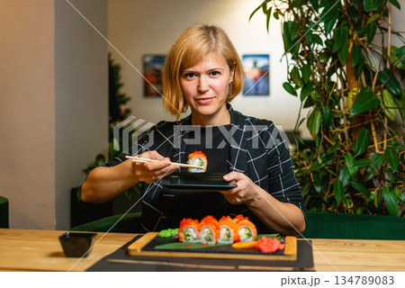 Woman smiles while lifting a sushi roll with chopsticks at a cozy restaurant table. Woman smiles while lifting a sushi roll with chopsticks at a cozy restaurant table. 134789083