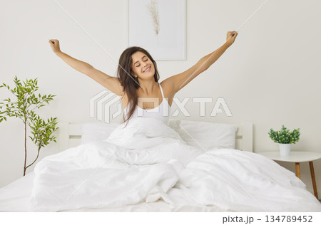 Happy caucasian young woman sitting in cozy bed, stretching after waking up in bright sunny bedroom. Happy caucasian young woman sitting in cozy bed, stretching after waking up in bright sunny bedroom. 134789452
