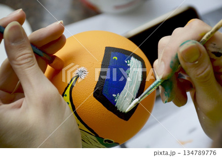 Female hands carefully painting a Christmas ball in vibrant orange hues Female hands carefully painting a Christmas ball in vibrant orange hues 134789776