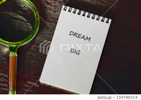 An open notebook with DREAM BIG inscribed lies beside a classic magnifying glass on a darkly stained wooden desk, alluding to grand ambitions and careful planning 134790314